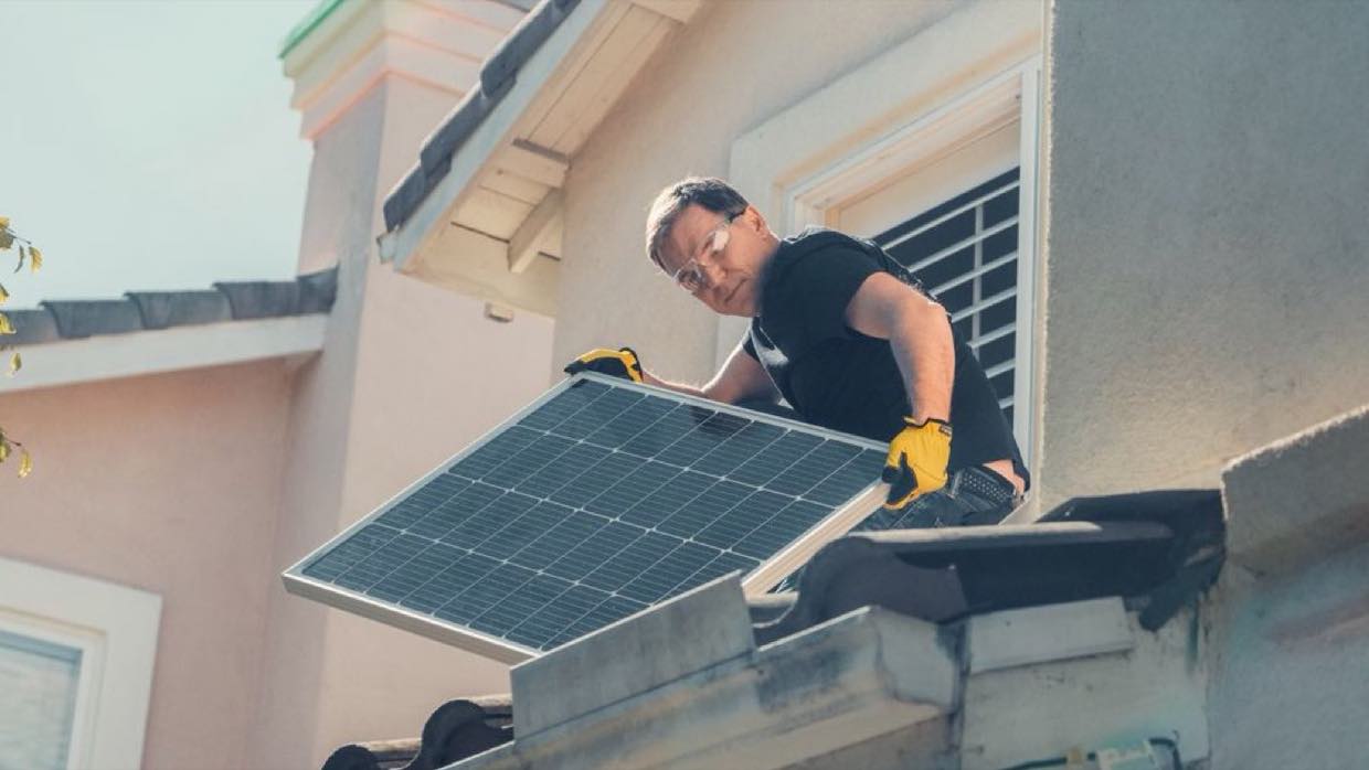 An installer in gloves guiding a solar panel into place at the edge of a roof.