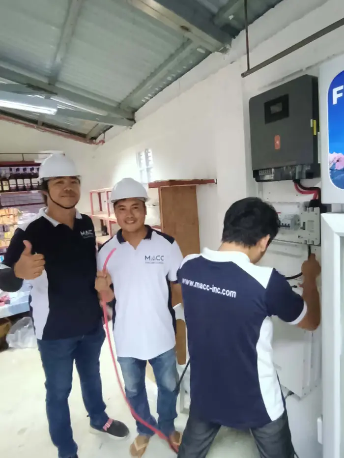 Three MACC team members in branded shirts and hard hats wiring the inverter indoors