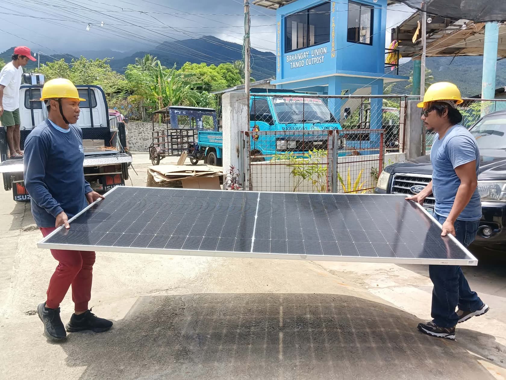 Two MACC installers carrying a solar panel past the Barangay Union Tanod Outpost in Caibiran